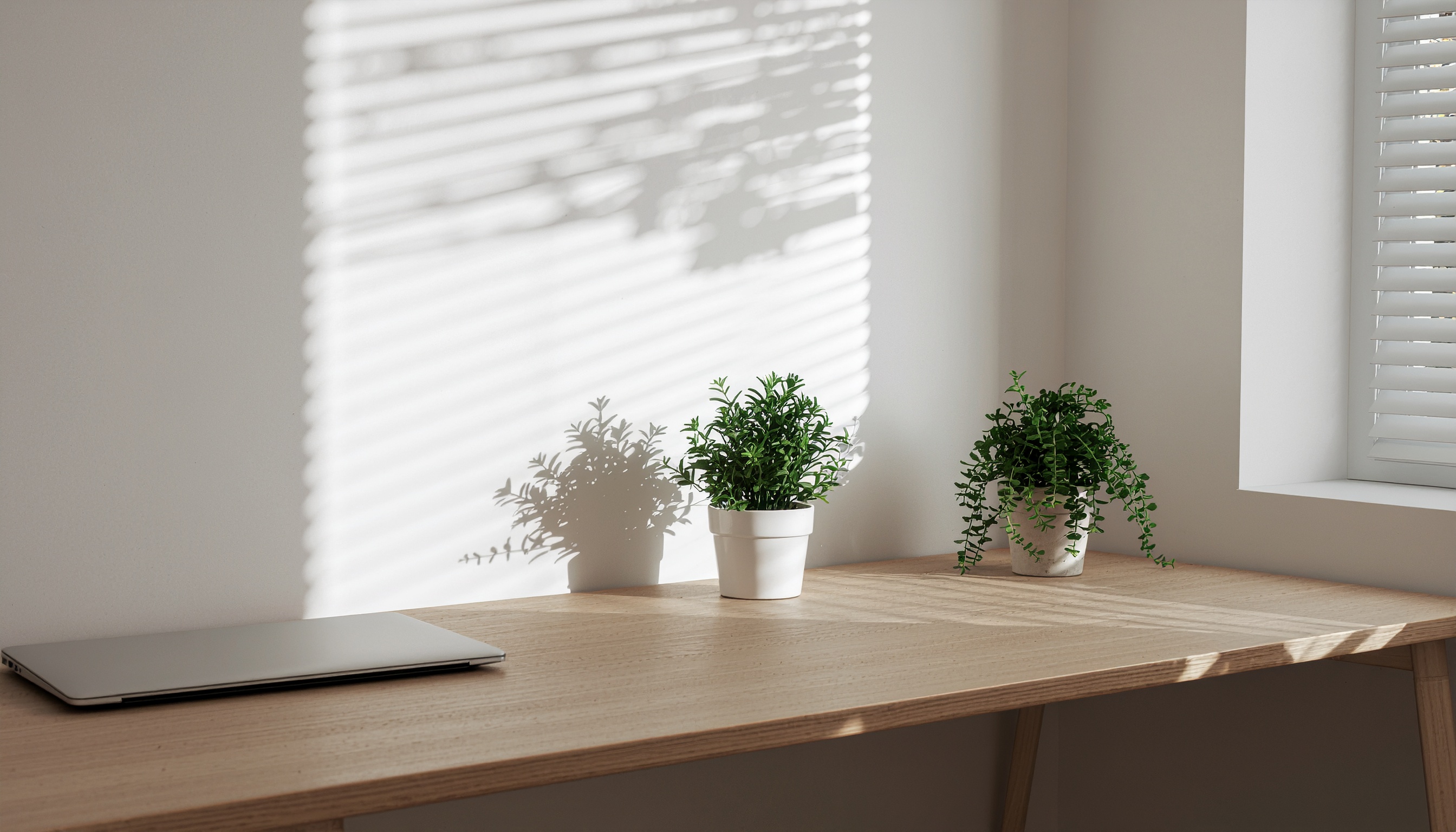 Cena minimalista de um espaço de trabalho moderno com mesa de madeira clara, dois vasos de plantas verdes e um laptop fechado sob luz suave. Persiana aberta projeta sombras lineares na parede, criando um efeito visual dinâmico e elegante. Composição equilibrada e serena, com foco em simplicidade e harmonia.